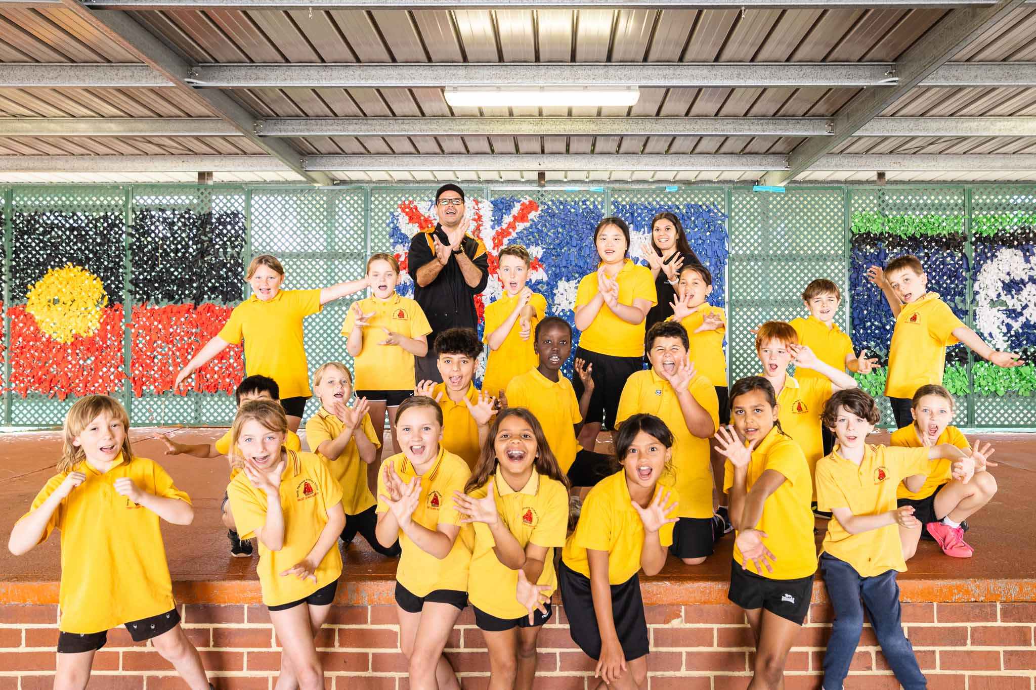 class of primary school students with teacher and teaching artist doing arts learning deadly arts lesson on indigenous dance in front of Australia, Aboriginal and Torres Strait Island flags