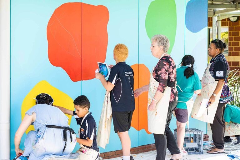 Teaching artist helping primary school students in arts learning visual arts lesson to paint bright indigenous themed school mural through their community project