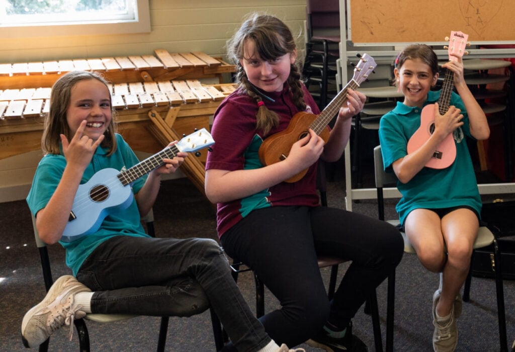 three smiling primary school students engaged in arts learning music lesson on ukuleles