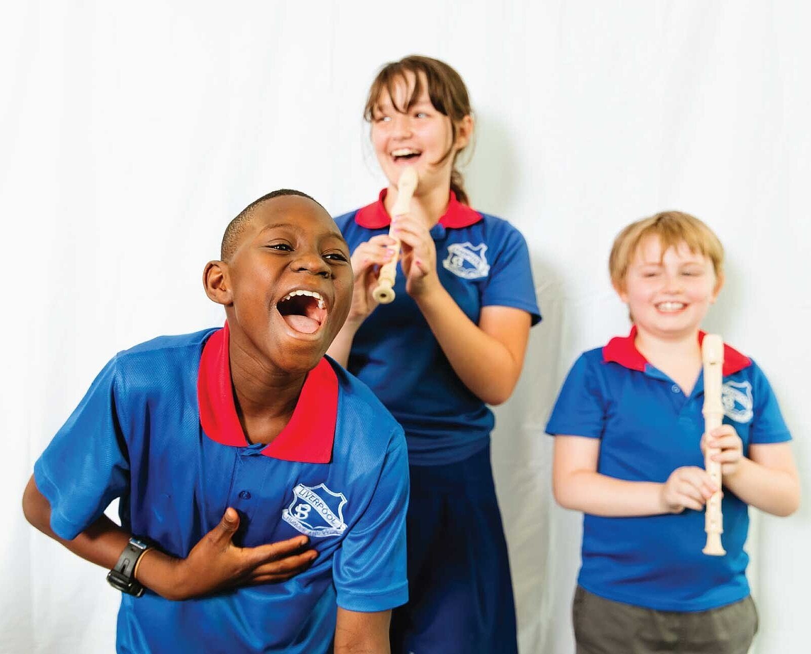 Three students in front of a white background. Two are playing the recorder and one is laughing to the camera.